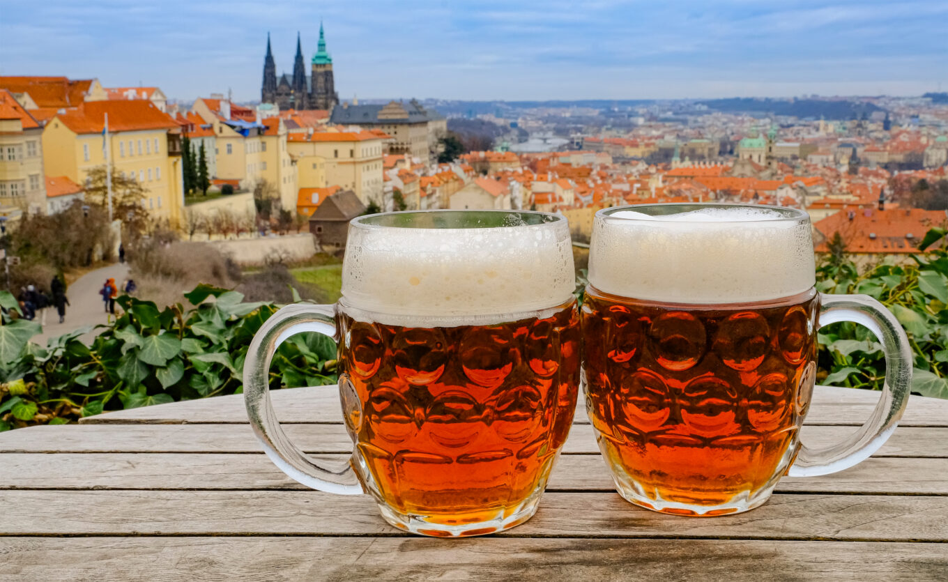 Zdjęcie przedstawia dwa kufle piwa na tle panoramy Pragi