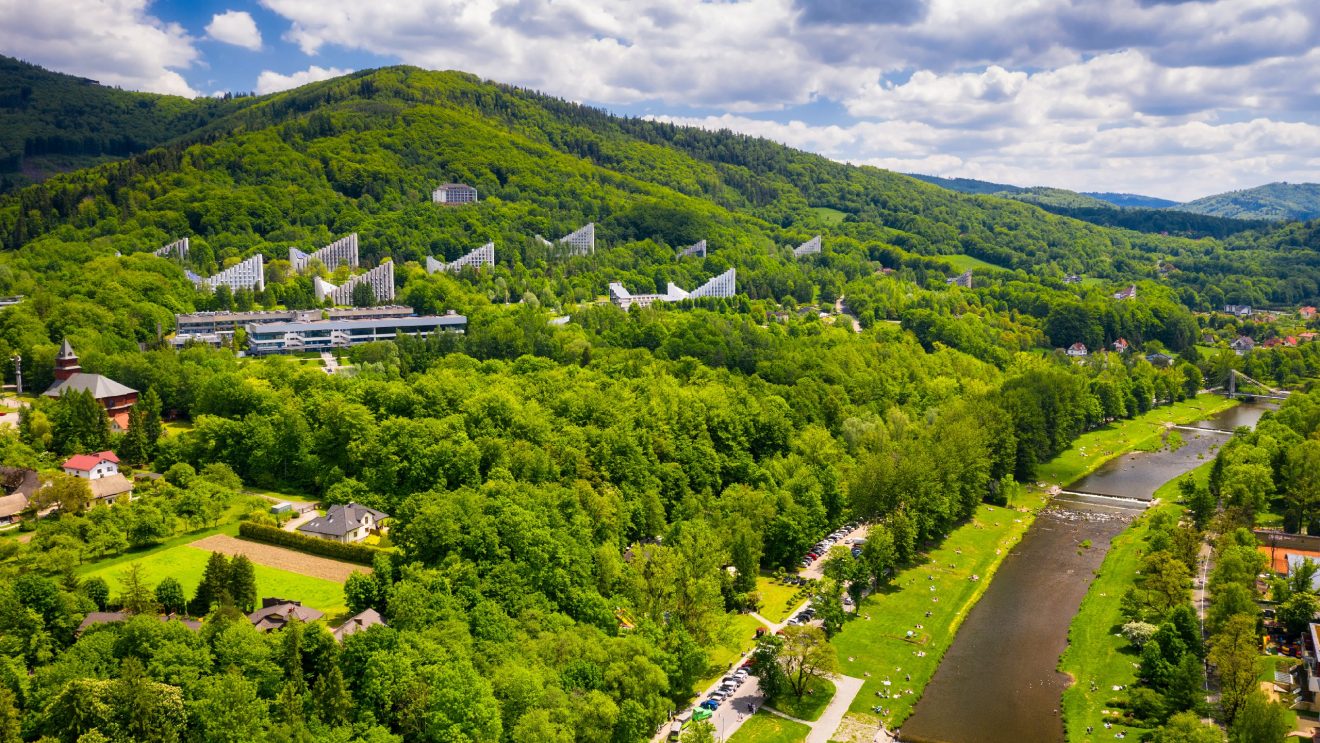Panorama Beskidów z wzgórzami porośniętymi lasem, pomiędzy drzewami budynki w kształcie trójkątów