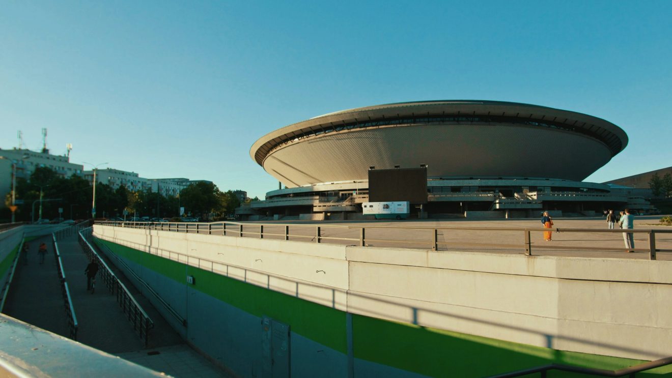 Nowoczesna hala widowiskowo-sportowa o futurystycznym kształcie, sfotografowana w słoneczny dzień.